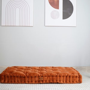 May include: A rectangular floor cushion in a burnt orange velvet fabric featuring a tufted design with multiple fabric-covered buttons across the top surface and a structured, boxy edge profile with a small rectangular brand label on the side.