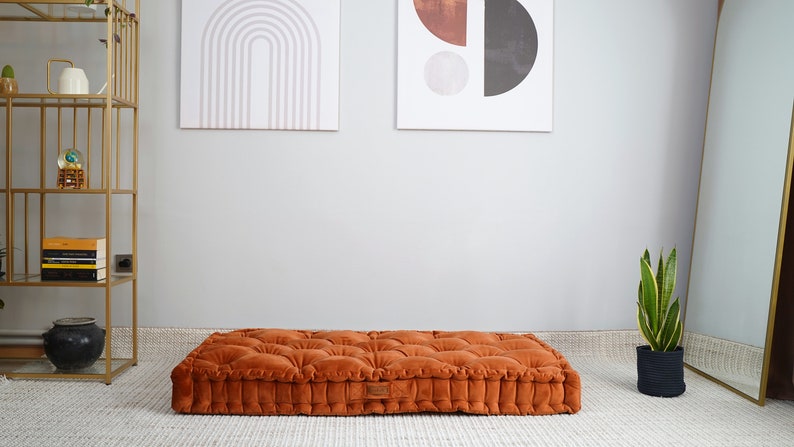May include: A rectangular floor cushion in a burnt orange velvet fabric featuring a tufted design with multiple fabric-covered buttons across the top surface and a structured, boxy edge profile with a small rectangular brand label on the side.