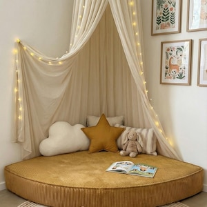 May include: A children's reading nook with a mustard yellow round floor cushion, a cream canopy with fairy lights, and decorative pillows. A wooden toy camera and stacking rings sit on a round rug.