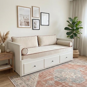 Può includere: Un divano letto bianco con tre cassetti, dotato di un materasso beige, due cuscini rettangolari per lo schienale e due cuscini cilindrici laterali, tutti rifiniti con un sottile bordino marrone lungo i profili del rivestimento.