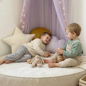 May include: Two young children are playing on a round, corduroy-covered floor cushion. One child is holding a wooden stacking toy, while the other is lying down with a stuffed bunny and wooden blocks. A lavender canopy with fairy lights hangs above.