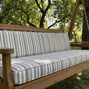 Op de afbeelding: Houten schommelbank met een gestreept kussen in blauw, beige en wit. De bank hangt aan een houten frame met metalen kettingen, in een tuin.