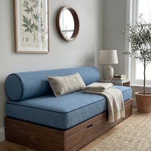 May include: A blue upholstered daybed with a wooden base and two drawers. A bolster pillow and decorative pillow rest on the bed. A beige throw blanket is draped over the bed. A framed botanical print and a round mirror hang on the wall.