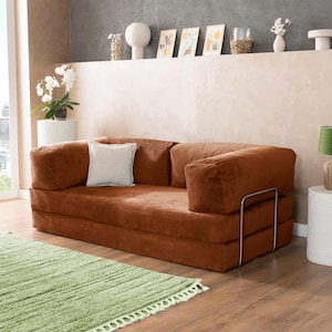 May include: A modern, corduroy-covered sofa in a warm brown hue, featuring a square shape and a minimalist metal frame. A white square pillow rests on the sofa. A light green rug with a fringe is in front of the sofa.