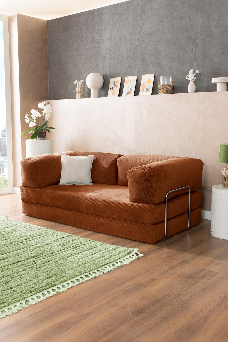 May include: A modern, corduroy-covered sofa in a warm brown hue, featuring a square shape and a minimalist metal frame. A white square pillow rests on the sofa. A light green rug with a fringe is in front of the sofa.