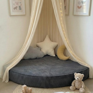 May include: A children's reading nook featuring a grey corduroy floor cushion, a cream canopy with fairy lights, and star, cloud, and crescent moon pillows. A teddy bear, wooden blocks, and a book are on a round jute rug. The room has framed pictures.