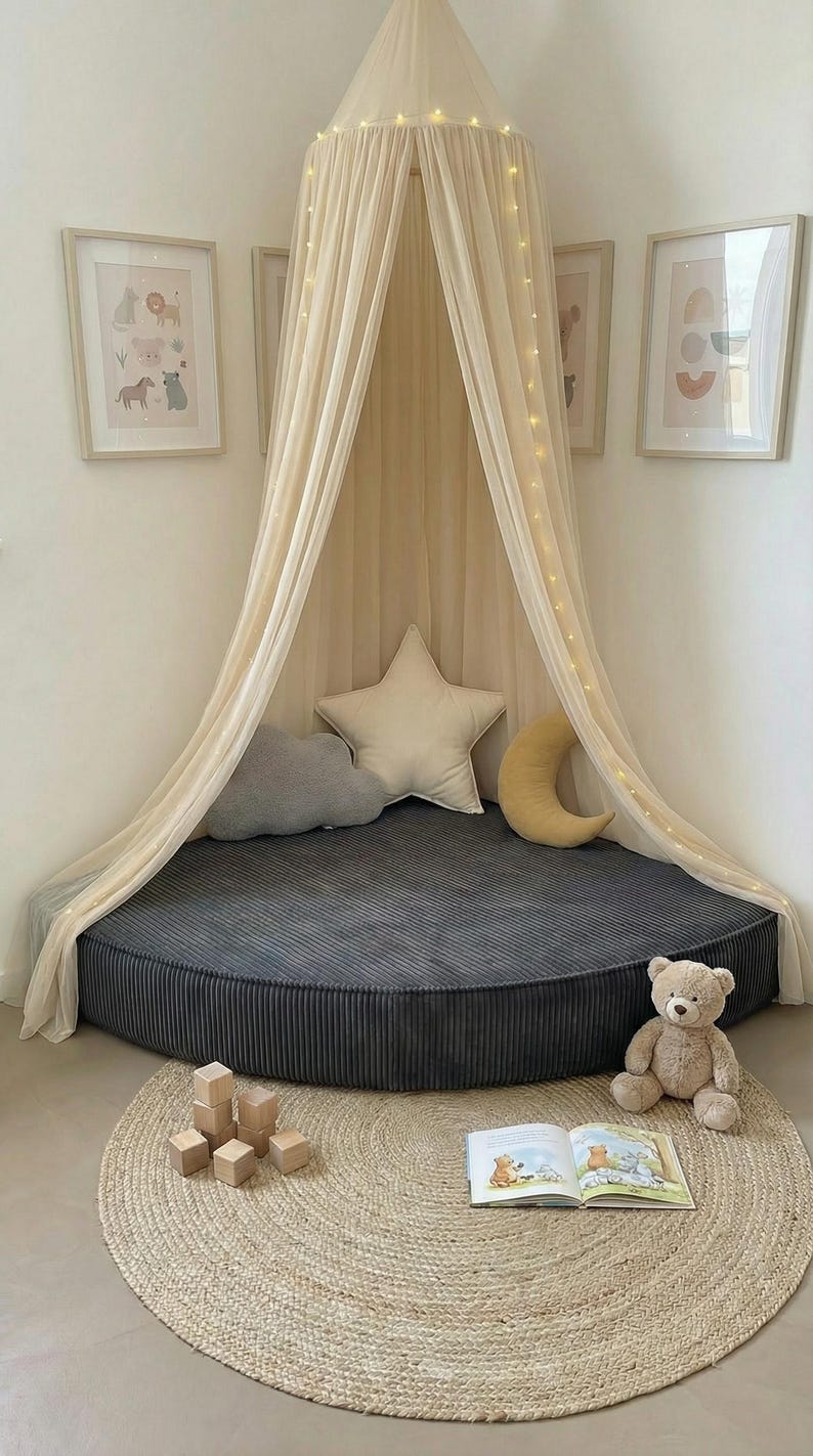 May include: A children's reading nook featuring a grey corduroy floor cushion, a cream canopy with fairy lights, and star, cloud, and crescent moon pillows. A teddy bear, wooden blocks, and a book are on a round jute rug. The room has framed pictures.