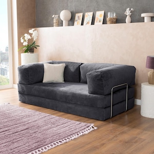 May include: A dark gray corduroy sofa with a square shape and a light-colored square pillow. The sofa has a metal frame and is placed on a wooden floor next to a purple rug. The room has a neutral color palette.
