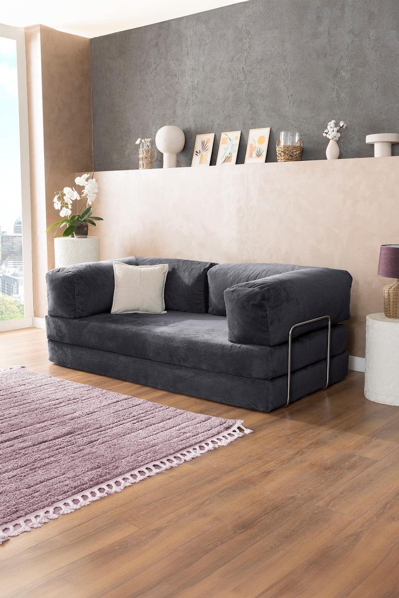 May include: A dark gray corduroy sofa with a square shape and a light-colored square pillow. The sofa has a metal frame and is placed on a wooden floor next to a purple rug. The room has a neutral color palette.