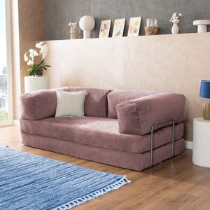 May include: A dusty rose corduroy sofa with a light-colored square pillow. The sofa has a metal frame and is placed on a wooden floor. A blue rug with a fringe is in front of the sofa. The room has a neutral color palette.
