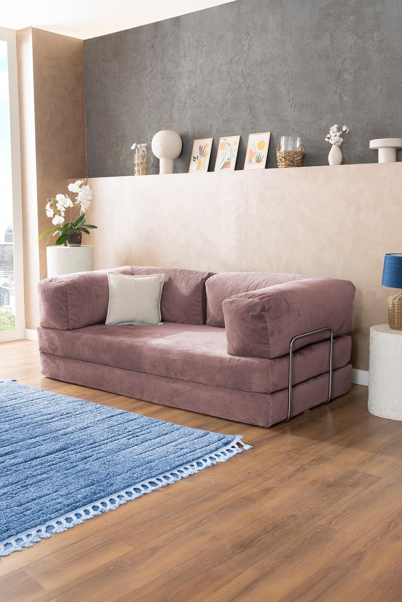 May include: A dusty rose corduroy sofa with a light-colored square pillow. The sofa has a metal frame and is placed on a wooden floor. A blue rug with a fringe is in front of the sofa. The room has a neutral color palette.