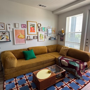 Op de afbeelding: Een mosterdkleurige hoekbank met een groen kussen en een patroon deken. Een blauw en rood geometrisch tapijt bedekt de vloer. De muur toont ingelijste kunstwerken en kleine planken. Een houten salontafel staat in het midden.