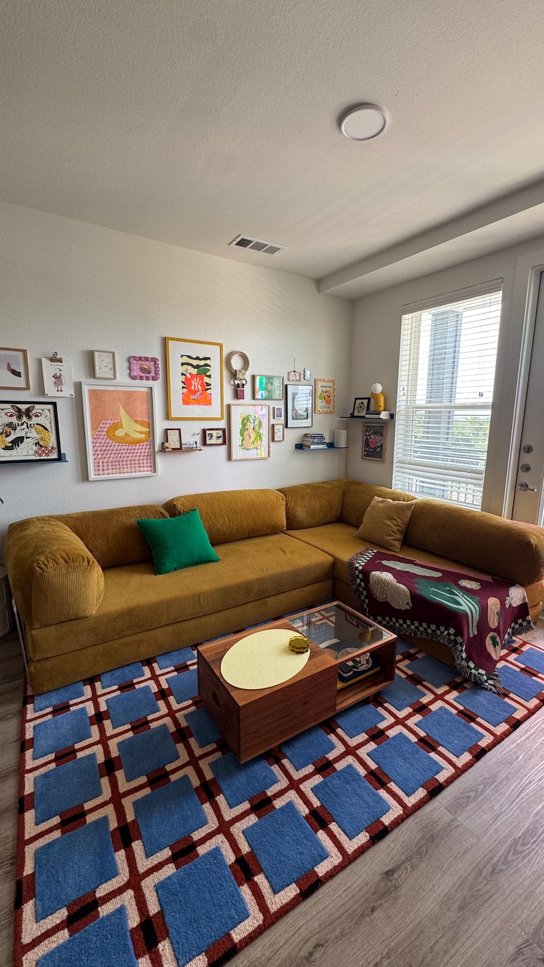 Op de afbeelding: Een mosterdkleurige hoekbank met een groen kussen en een patroon deken. Een blauw en rood geometrisch tapijt bedekt de vloer. De muur toont ingelijste kunstwerken en kleine planken. Een houten salontafel staat in het midden.