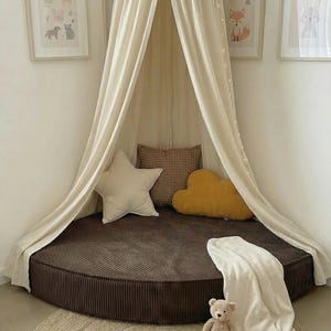 May include: A children's reading nook featuring a brown corduroy bed, cream canopy, and decorative pillows. A jute rug, teddy bear, and framed animal art complete the space. Fairy lights provide a soft glow.