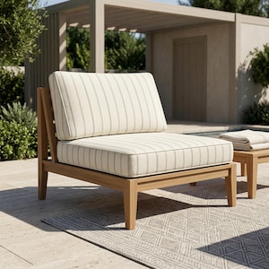 May include: A wooden outdoor armchair with a cream-colored cushion featuring vertical gray stripes. The chair has a low back and a matching cushion. A small side table with a folded blanket is next to the chair. The furniture is on a stone patio.