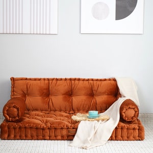 May include: A low-profile, rust-colored velvet floor sofa featuring a tufted design with a rectangular seat, a backrest, and two cylindrical bolster armrests. A cream-colored throw blanket is draped over the right side of the sofa.
