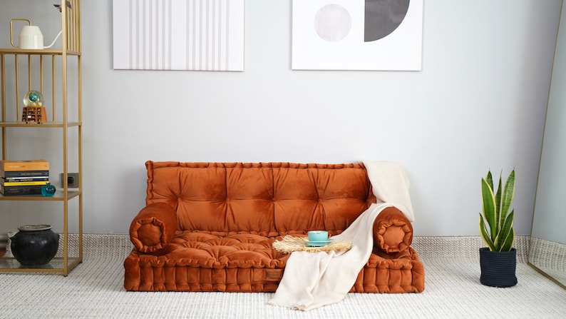 May include: A low-profile, rust-colored velvet floor sofa featuring a tufted design with a rectangular seat, a backrest, and two cylindrical bolster armrests. A cream-colored throw blanket is draped over the right side of the sofa.