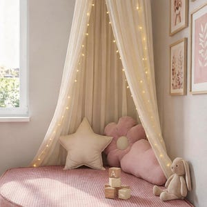 May include: A child's reading nook featuring a pink corduroy bed, star and flower-shaped cushions, and a cream canopy with fairy lights. Framed artwork and a stuffed bunny create a welcoming atmosphere. The bed is approximately 150 cm in diameter.