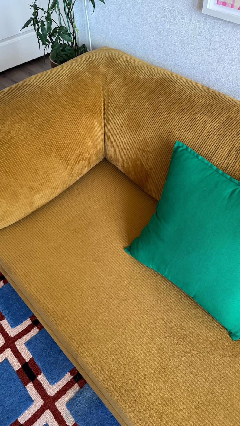 Op de afbeelding: Mosterdgeel corduroy bank met een groen kussen. De bank heeft een geribbelde textuur en een modern design. Een kleurrijk tapijt met een geometrisch patroon is zichtbaar. De bank is een stijlvol meubelstuk.