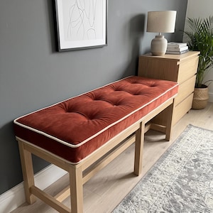 May include: A rectangular bench featuring a tufted rust-colored velvet cushion with white piping trim, supported by a light-toned wooden frame with four straight legs and horizontal support bars connecting the legs on each side.