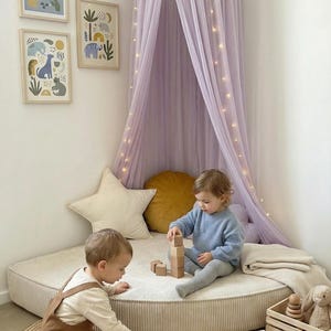 May include: A nursery scene with two children playing. A lilac canopy with fairy lights is suspended above a circular play mat. The room includes framed animal prints, a star cushion, and a wooden toy train.