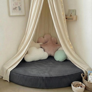 May include: A children's reading nook with a cream canopy and fairy lights. The circular bed has a dark grey corduroy cover and decorative pillows shaped like a cloud, star, and flower. A framed animal print is on the wall. The bed is approximately 150 cm in diameter.