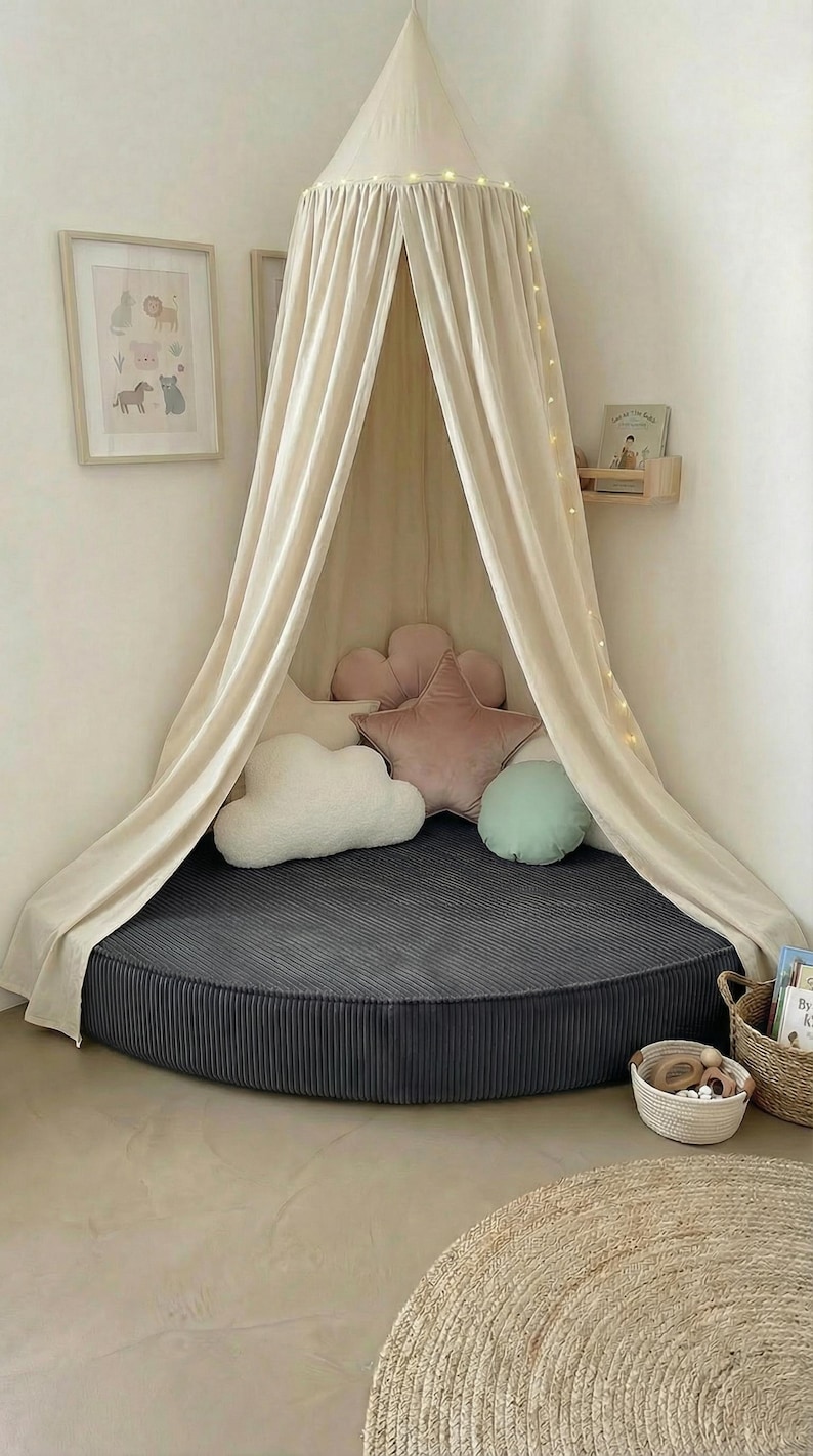 May include: A children's reading nook with a cream canopy and fairy lights. The circular bed has a dark grey corduroy cover and decorative pillows shaped like a cloud, star, and flower. A framed animal print is on the wall. The bed is approximately 150 cm in diameter.