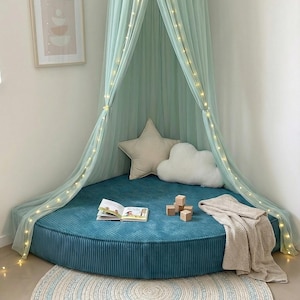 May include: A teal corduroy floor cushion with a light green canopy and fairy lights creates a cosy reading nook. Includes star and cloud cushions, a blanket, a book, wooden blocks, and a round rug with blue accents.