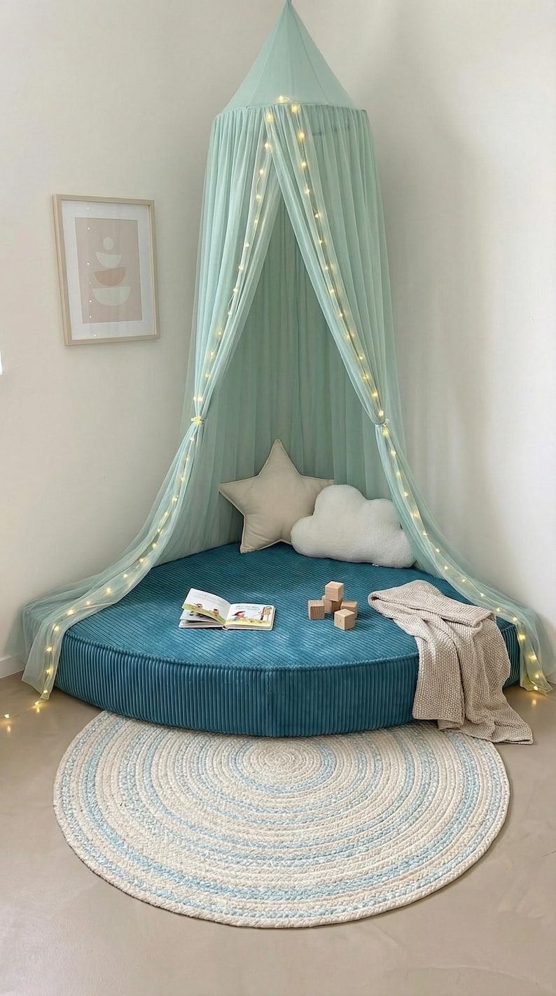 May include: A teal corduroy floor cushion with a light green canopy and fairy lights creates a cosy reading nook. Includes star and cloud cushions, a blanket, a book, wooden blocks, and a round rug with blue accents.