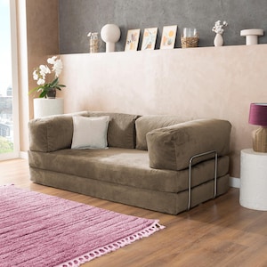May include: A modern, low-profile sofa in a neutral brown corduroy fabric. The sofa has a square shape with large, cushioned armrests and a matching back. A small, square white pillow rests on the sofa. A pink rug is in front of the sofa.