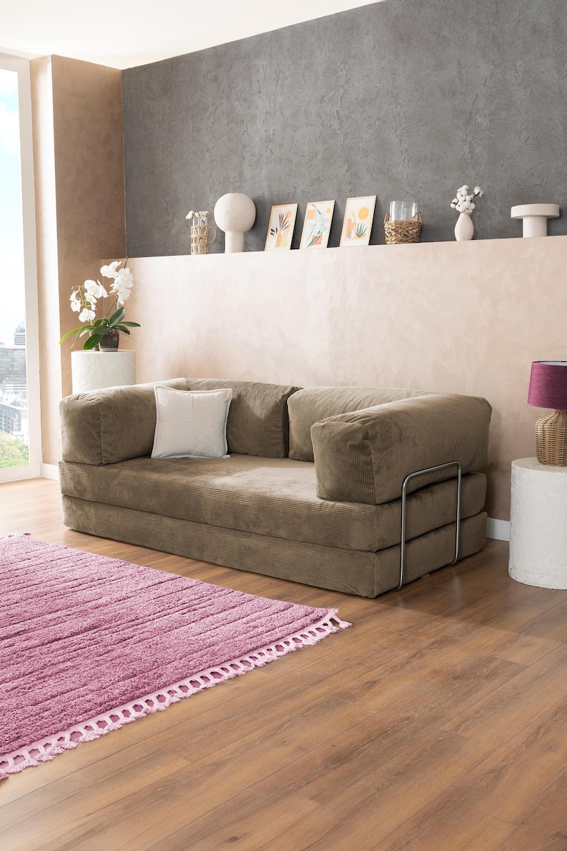 May include: A modern, low-profile sofa in a neutral brown corduroy fabric. The sofa has a square shape with large, cushioned armrests and a matching back. A small, square white pillow rests on the sofa. A pink rug is in front of the sofa.