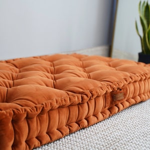 May include: A rectangular floor cushion upholstered in a burnt orange velvet fabric. The cushion features a tufted surface with multiple indentations and a thick, structured border with vertical pleats. A small rectangular brand label is attached.