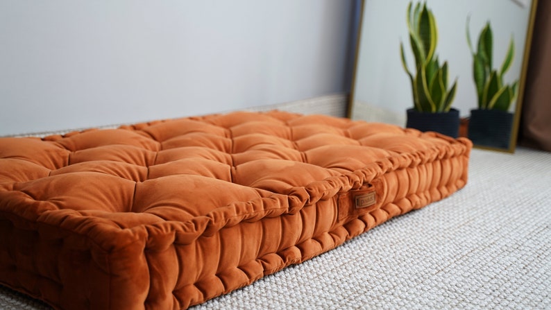 May include: A rectangular floor cushion upholstered in a burnt orange velvet fabric. The cushion features a tufted surface with multiple indentations and a thick, structured border with vertical pleats. A small rectangular brand label is attached.