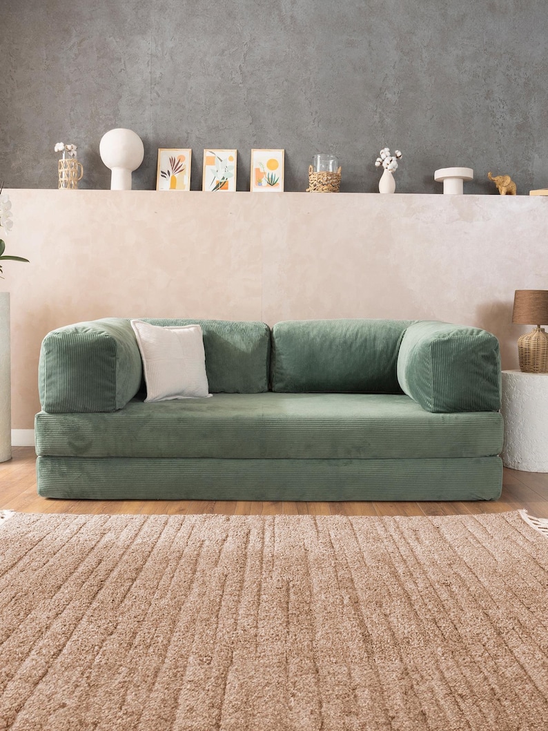 May include: A green corduroy sofa with a matching corduroy base and armrests. A white square pillow rests on the sofa. The sofa sits on a light brown rug. Decorative items are on a shelf in the background.
