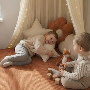 May include: Two children playing on a round, rust-coloured corduroy mat. One child is lying down with a star-shaped pillow, while the other sits with wooden blocks and a stuffed fox. The scene is set under a draped canopy with string lights.