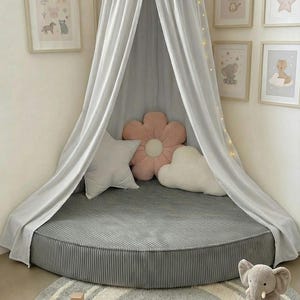 May include: A grey, circular children's play area with a canopy, pillows, and a rug. The canopy drapes over a circular bed. Framed animal prints decorate the wall. A plush elephant toy and wooden blocks are on the rug.