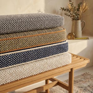 May include: A stack of four rectangular bench cushions with a textured, woven fabric finish. The cushions are colored in black, mustard yellow, navy blue, and cream, each featuring a contrasting piped edge detail along the side seams.