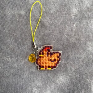 Peut inclure: Un petit charme en forme de poulet pixelis&eacute; orange et jaune, suspendu &agrave; un cordon jaune. Le charme est orn&eacute; d'une perle transparente de couleur ambre.