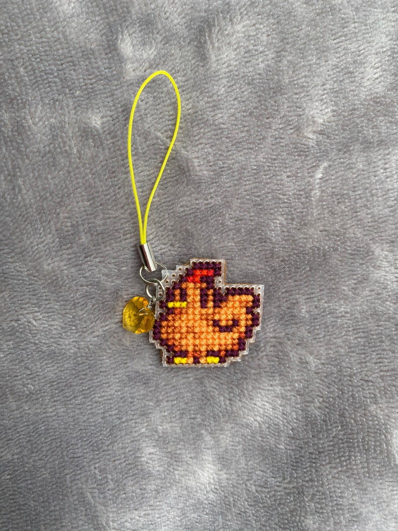 Peut inclure: Un petit charme en forme de poulet pixelis&eacute; orange et jaune, suspendu &agrave; un cordon jaune. Le charme est orn&eacute; d'une perle transparente de couleur ambre.