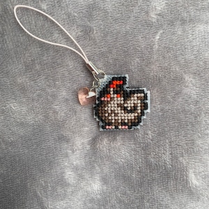 Peut inclure: Un porte-cl&eacute;s en cordon blanc avec un petit charme en forme de c&oelig;ur rose et un motif de point de croix repr&eacute;sentant un poulet brun avec une cr&ecirc;te rouge.