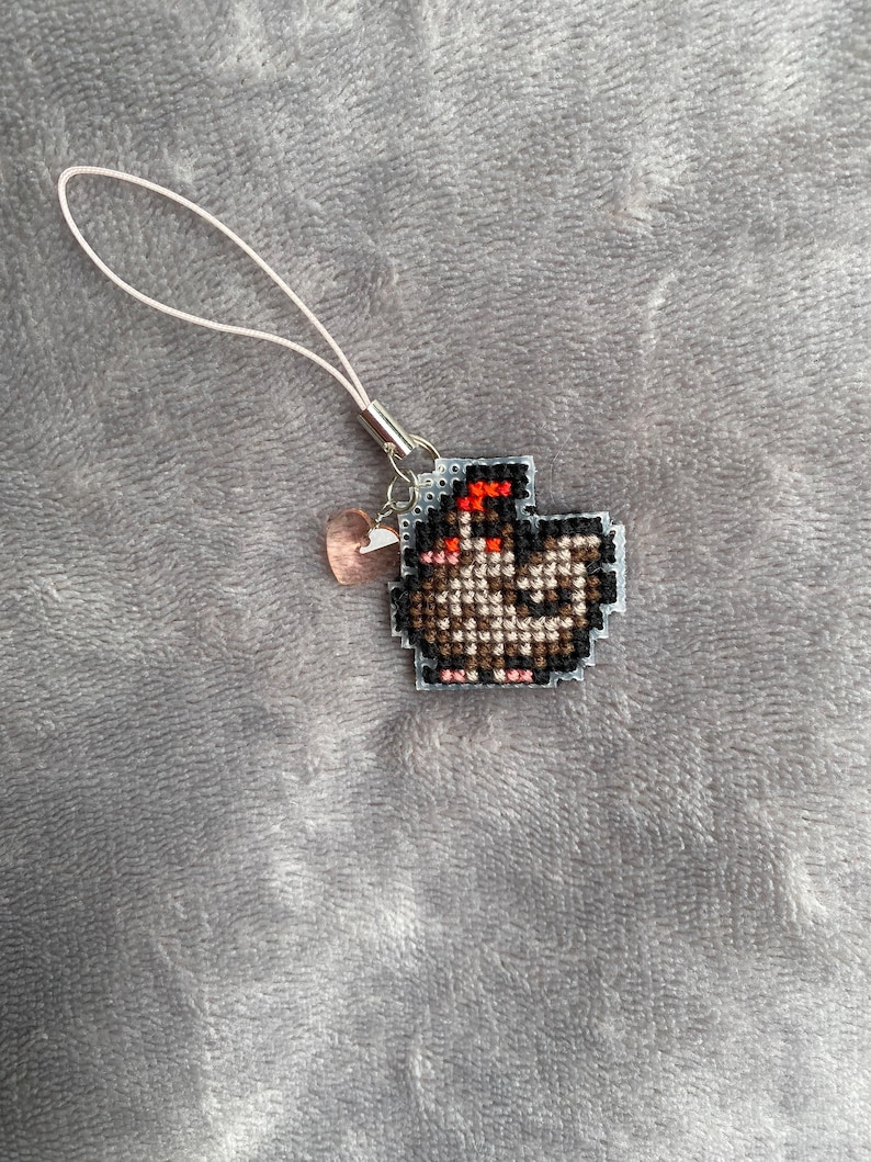 Peut inclure: Un porte-cl&eacute;s en cordon blanc avec un petit charme en forme de c&oelig;ur rose et un motif de point de croix repr&eacute;sentant un poulet brun avec une cr&ecirc;te rouge.
