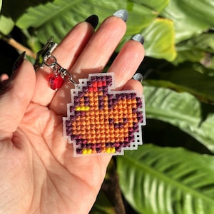 Peut inclure: Un petit porte-cl&eacute;s en point de croix marron et orange repr&eacute;sentant un poulet de dessin anim&eacute;. Le porte-cl&eacute;s est attach&eacute; &agrave; une cha&icirc;ne argent&eacute;e avec une perle rouge.
