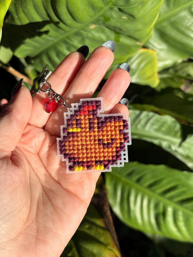 Peut inclure: Un petit porte-cl&eacute;s en point de croix marron et orange repr&eacute;sentant un poulet de dessin anim&eacute;. Le porte-cl&eacute;s est attach&eacute; &agrave; une cha&icirc;ne argent&eacute;e avec une perle rouge.