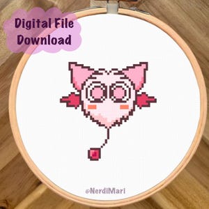 May include: A pixelated pink creature cross-stitch design in a wooden embroidery hoop. The design features a heart-shaped body, large eyes, and small wings. Text in a pink bubble reads "Digital File Download". The artist's name, @NerdiMari, is at the bottom.