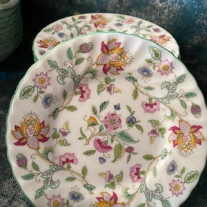 Puede incluir: Un juego de platos de cerámica blanca con un borde verde y un patrón floral en tonos de rosa, amarillo y verde. El patrón presenta flores y hojas estilizadas.