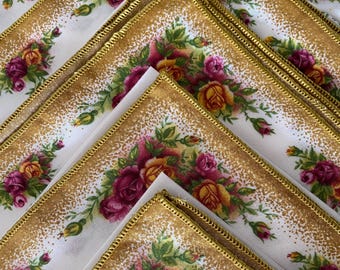 May include: A set of white linen napkins with a gold trim and a floral pattern featuring pink and yellow roses.