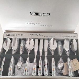 May include: A set of stainless steel cutlery in a box with the words "Monogram Old Country Roses Royal Albert" printed on the box. The cutlery has a floral design on the handles.