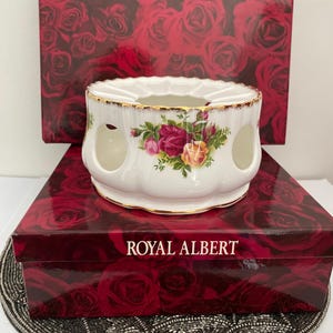 Może przedstawiać: Biała popielniczka Royal Albert z kwiatowym wzorem różowych, żółtych i czerwonych róż, z akcentami ze złotych zdobień. Popielniczka stoi na czerwonym pudełku z wzorem róż i napisem "ROYAL ALBERT".