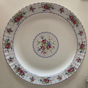 Puede incluir: Un plato de cerámica blanco con un patrón floral azul y morado. El plato tiene un borde festoneado y un diseño floral central.
