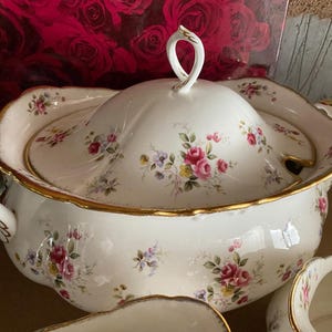 Puede incluir: Un tazón de servir de porcelana blanca con tapa, con un patrón floral con rosas rosadas y ribetes dorados. El tazón está decorado con un delicado diseño floral.